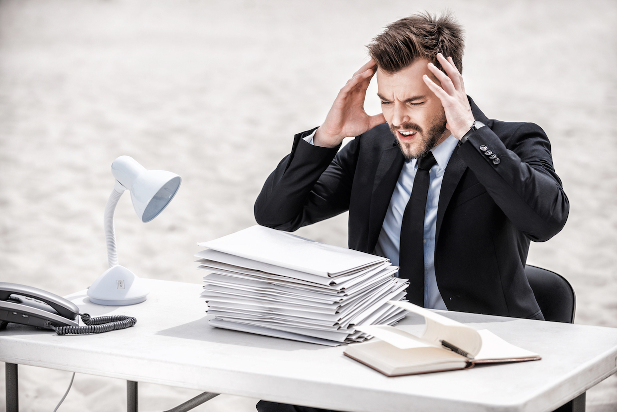 No more paperwork! Frustrated young businessman holding head in hands while sitting at the table on sand